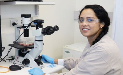 Dr Taskeen Janjua is targeting cancer cells with a drug delivered by a new nanoparticle. a woman with dark hair wearing safety glasses sits in front of a white and black microscope